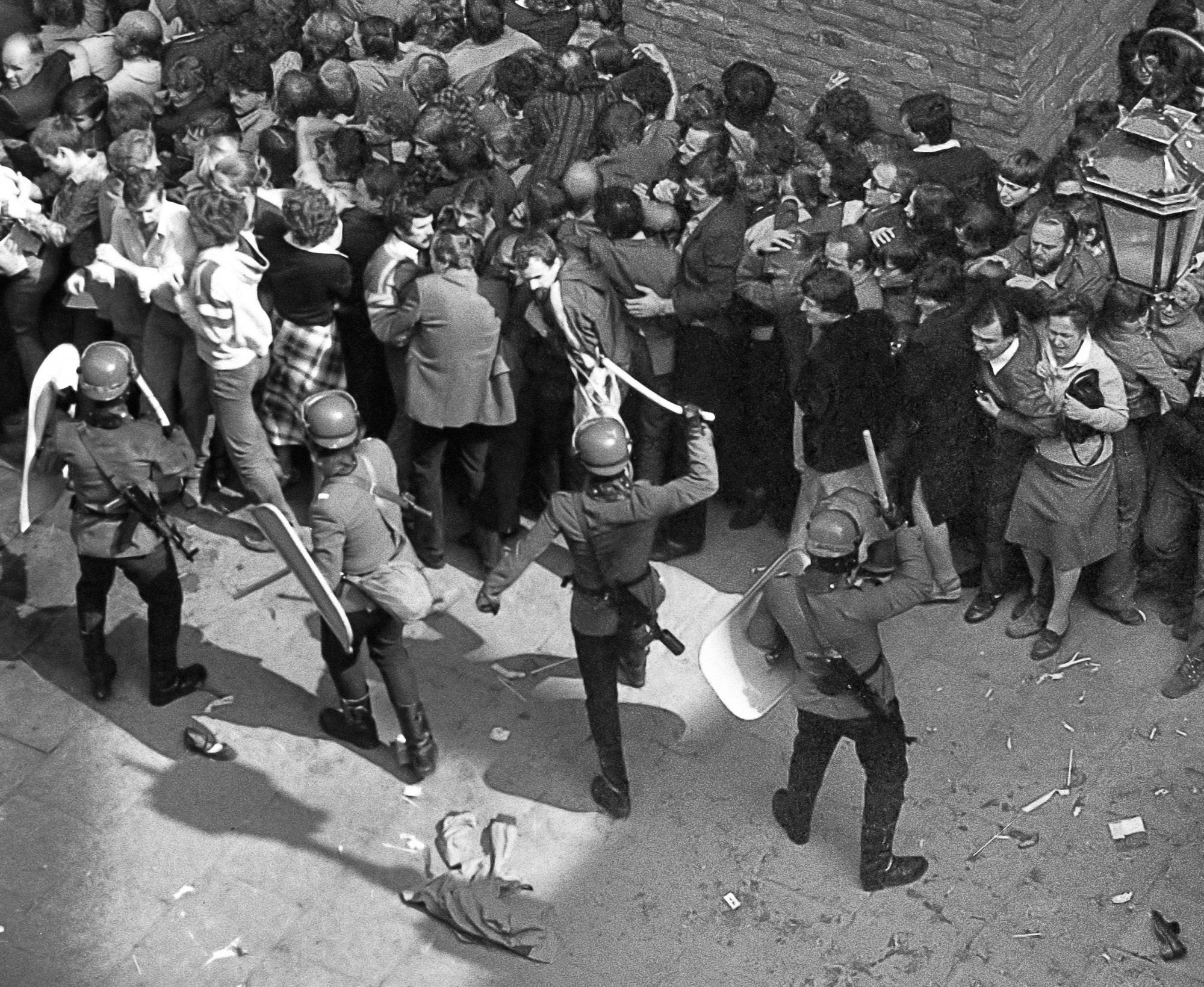Czarno-białe zdjęcie przedstawiające starcie umundurowanych policjantów w hełmach, z tarczami i pałkami, z gęstym tłumem cywilów na ulicy podczas protestu.